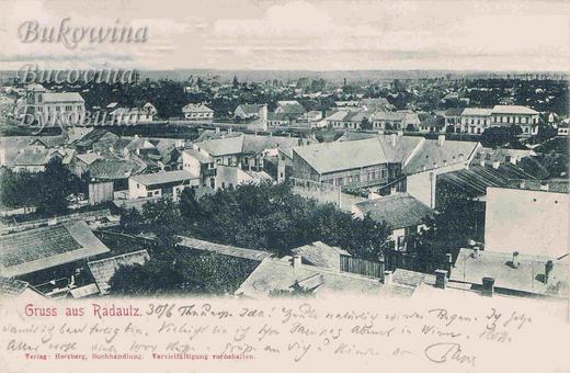 View over tiled rooftops and gardens to the flat town beyond, with a large twin-towered building at left; handwritten note