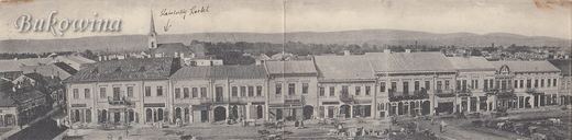 Panorama of a row of two-storey shops on the main square with carts in front; an arrow marks a church tower behind