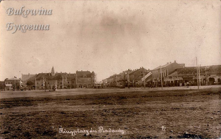 A wide empty square of bare earth with a row of two-storey buildings and a domed tower in the distance