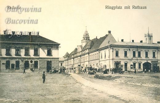 Square with a man standing in the foreground, a corner building and the town hall with a domed tower and parked carriages