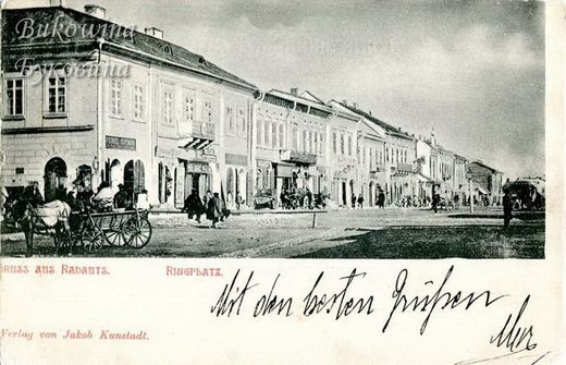 Row of two-storey shops with signboards along the square, a horse cart in front; handwritten greeting below