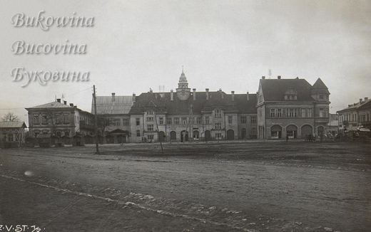 Broad empty square before the town hall with its domed clock tower, flanked by shops and wooden booths
