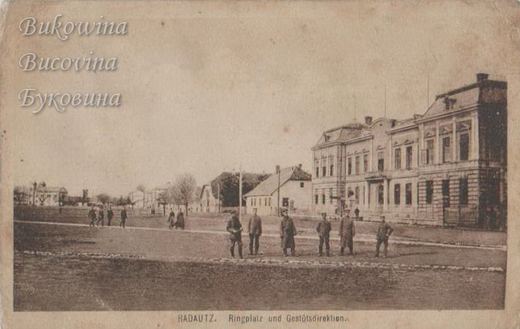 Wide open square with six uniformed soldiers standing in a row and a large two-storey building on the right