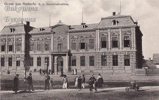 Stately two-storey building with a mansard roof and a pediment, people strolling in front, one with a parasol