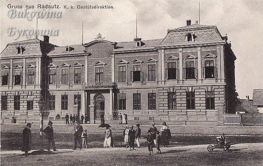 Stately two-storey building with a mansard roof and a pediment, people strolling in front, one with a parasol