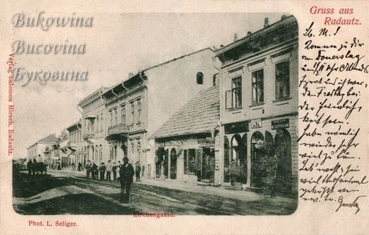 Unpaved street of two-storey shops with arched doors, men in hats on the pavement and a horse cart far off