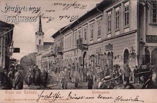 Busy street crowded with men in hats, a long row of buildings with shops and a church tower at the end