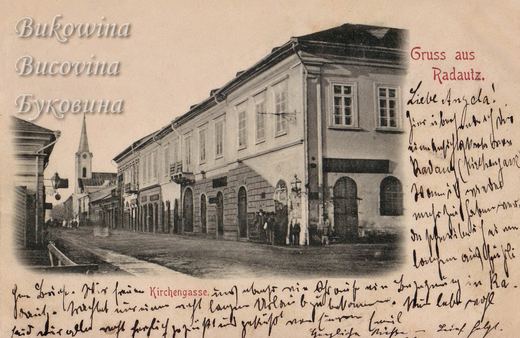 Quiet street with a long corner building with arched doors, a few people and a church spire in the distance