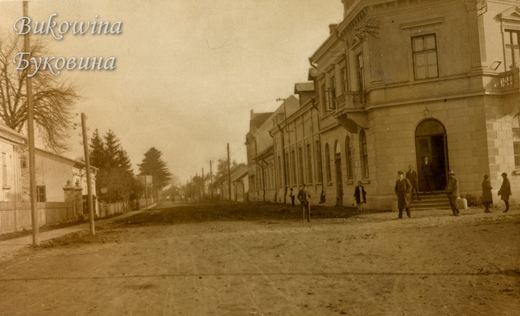 Wide unpaved street with a large corner building, men in coats on the steps and fir trees on the left