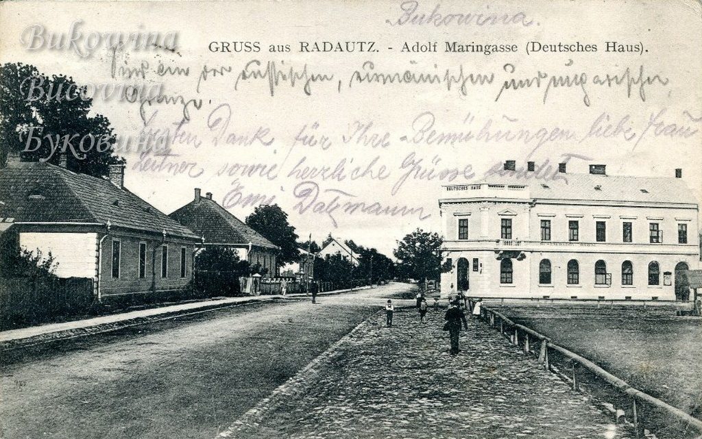 Cobbled path beside a wide road with low houses on the left and a large white building on the right