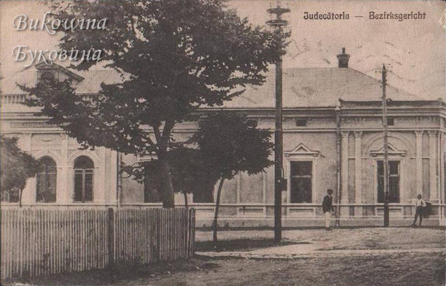 Single-storey building with arched windows partly hidden by a large tree and a wooden fence, two men in front