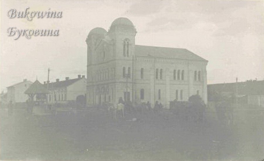 Large twin-towered temple with domed towers and arched windows beside low houses, an open square in front