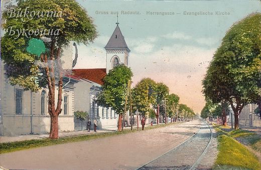 Coloured view of a tree-lined street with rail track, the Lutheran church tower and red-roofed houses at left