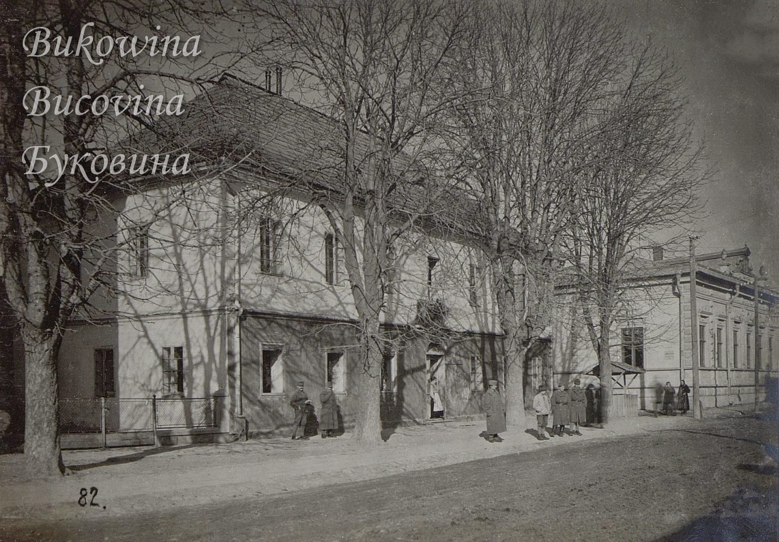 Two-storey house behind bare trees on a sunlit street, soldiers in greatcoats standing on the pavement