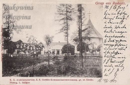 Garden with tall trees before low buildings and a church with a high conical roof, handwritten note at right