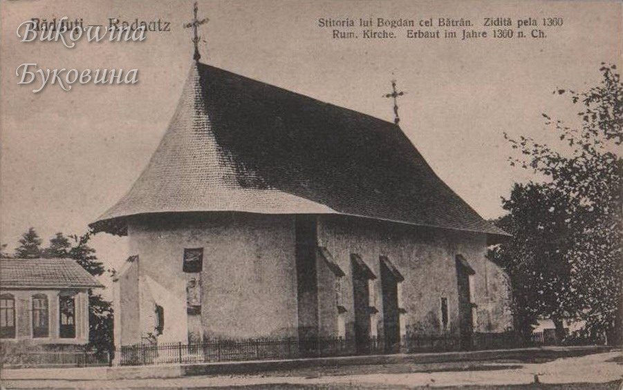 Old stone church with buttresses and a huge steep shingled roof topped by crosses, behind an iron fence