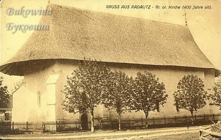 Old white church with a massive steep shingled roof, a row of young trees and an iron fence in front