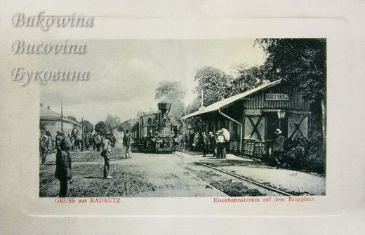 Small steam train at a wooden station signed Radautz-Ringplatz, crowds and a woman with a parasol nearby