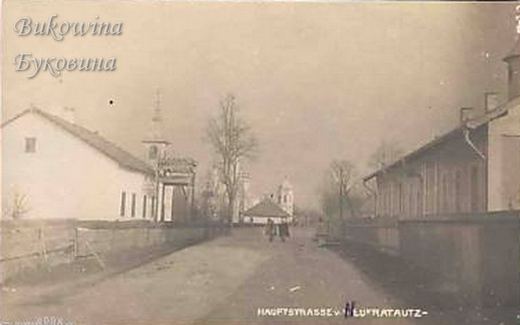 Unpaved village street between fences and houses, church towers in the distance and a few people