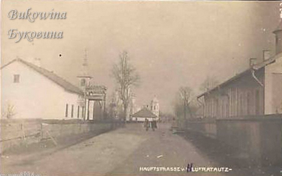 Unpaved village street between fences and houses, church towers in the distance and a few people