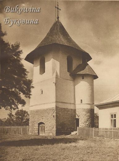 Stone and white-plastered bell tower with a shingled roof, a round stair turret and a cross, behind a fence