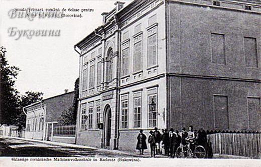 Two-storey corner school building on a street, a group of people and a man with a bicycle in front of the fence