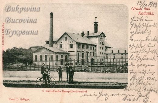 Brewery with a tall chimney behind an iron fence, men and a boy with a bicycle in front, handwritten message at right