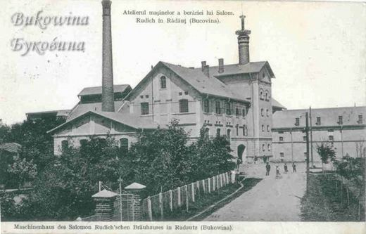 Brewery engine house with a tall chimney and tower, a lane with people walking and trees behind a fence