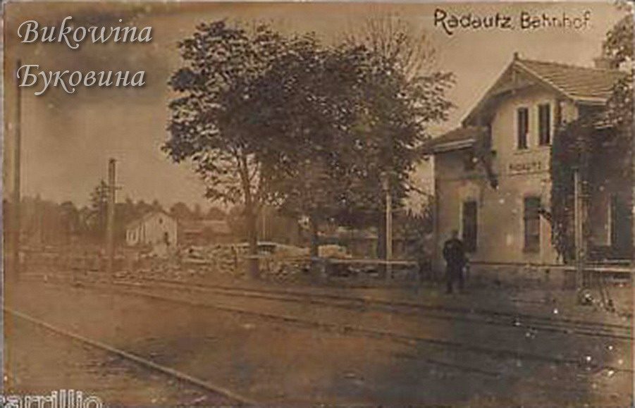 Small two-storey station building beside railway tracks, a man standing on the platform and trees beyond