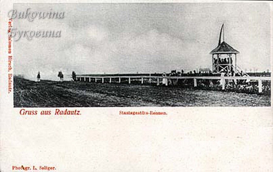 Racecourse with a white rail fence, spectators and a raised pavilion with a pennant, riders in the distance