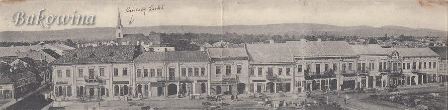 Panorama of a row of two-storey shops on the main square with carts in front; an arrow marks a church tower behind