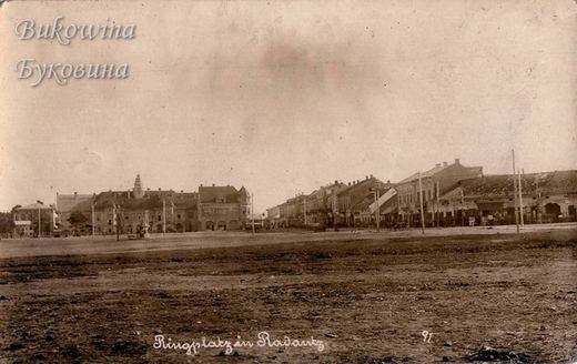 A wide empty square of bare earth with a row of two-storey buildings and a domed tower in the distance