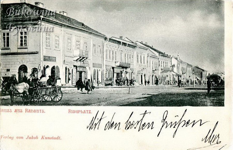 Row of two-storey shops with signboards along the square, a horse cart in front; handwritten greeting below