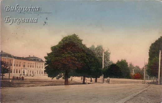 Tinted view of a broad square with large trees and a long pink two-storey building at left