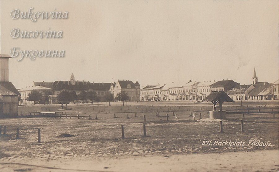 Open market square with wooden posts and a roofed well, a row of buildings and a church spire beyond