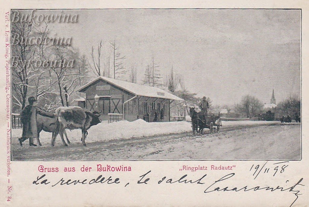 Snowy square with a timber pavilion, a man leading a cow, a horse cart and a church spire in the distance