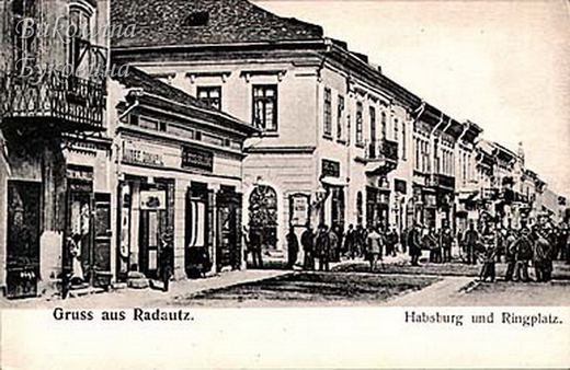 Corner of two-storey shop buildings with signs, and a crowd of men standing on the square in front