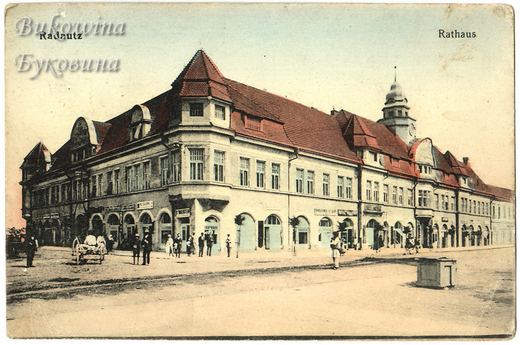 Hand-coloured view of a long red-roofed town hall with a clock tower, arched shopfronts and people on the square
