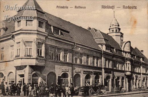 Town hall with a domed clock tower, a crowd of people and a cart stacked with timber in front of the shops