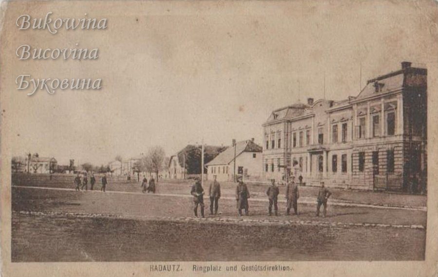 Wide open square with six uniformed soldiers standing in a row and a large two-storey building on the right
