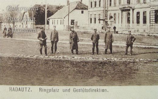 Close view of six uniformed soldiers standing on a square, with a house and a large building behind them