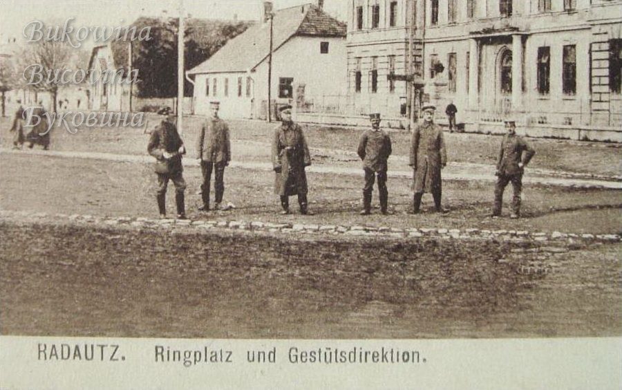 Close view of six uniformed soldiers standing on a square, with a house and a large building behind them