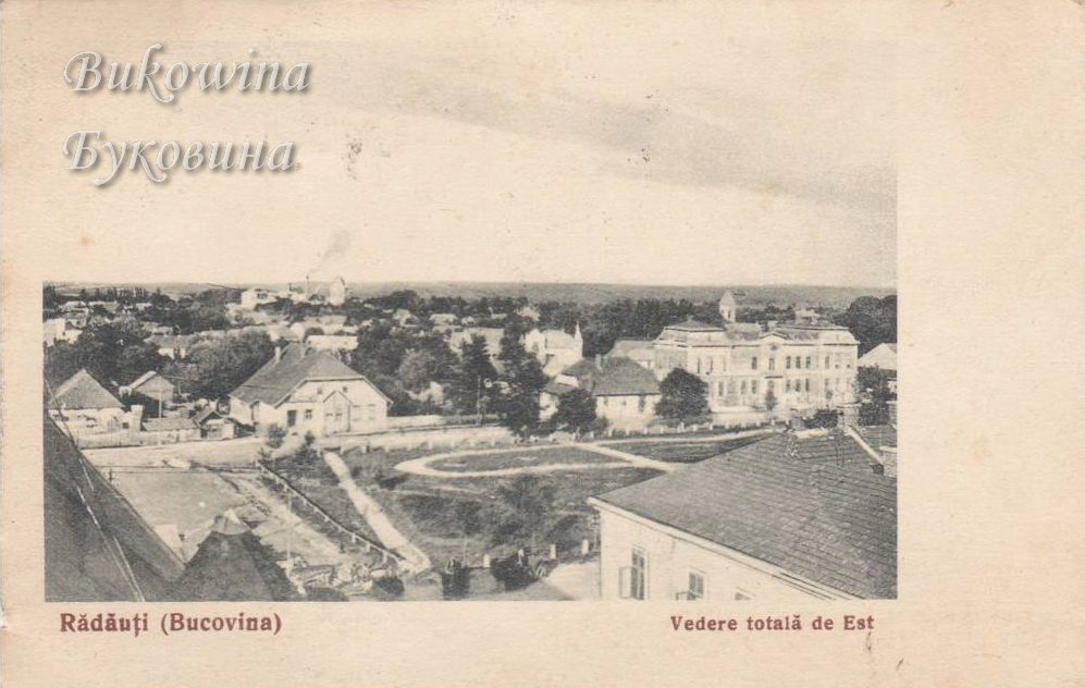 View over rooftops, gardens and a curving path towards a large two-storey building, with flat country beyond