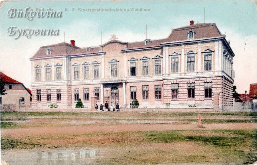 Coloured view of a pink and cream building with a mansard roof, a few people at the gate and a grassy square