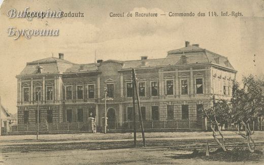 Two-storey building with a mansard roof behind iron railings, with telegraph poles and young trees in front