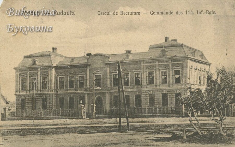 Two-storey building with a mansard roof behind iron railings, with telegraph poles and young trees in front