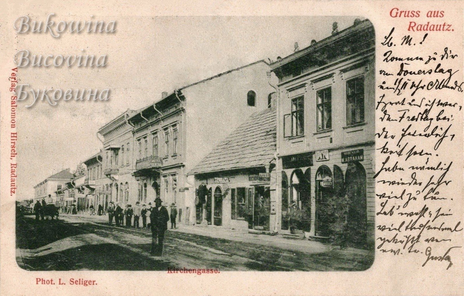 Unpaved street of two-storey shops with arched doors, men in hats on the pavement and a horse cart far off