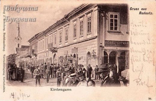 Crowded street of men in hats beside a long building with the shop sign Feibel Gutman, a church tower behind