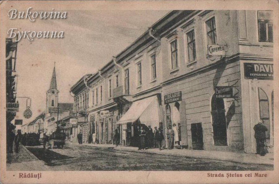 Street of two-storey buildings with a bookshop sign and an awning, people on the pavement and a church spire