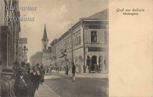Street with Hotel Bristol and the shop sign Feibel Gutman on a corner building, pedestrians and a church spire
